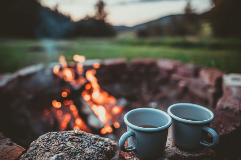 two mugs of coffee sitting next to a campfire.