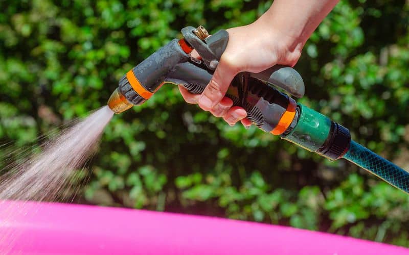 A hand is spraying a pink pool with a garden hose.