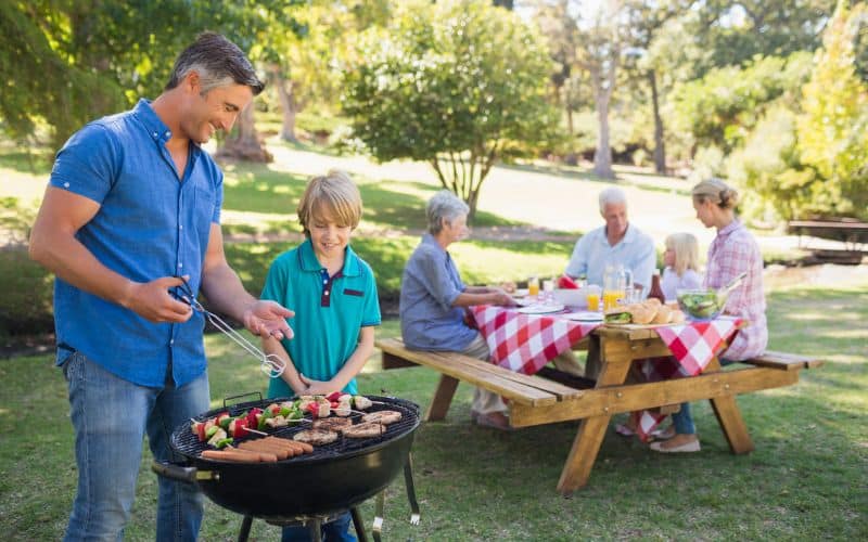 A family is grilling hamburgers on a grill and eating on a pedestal picnic table.
